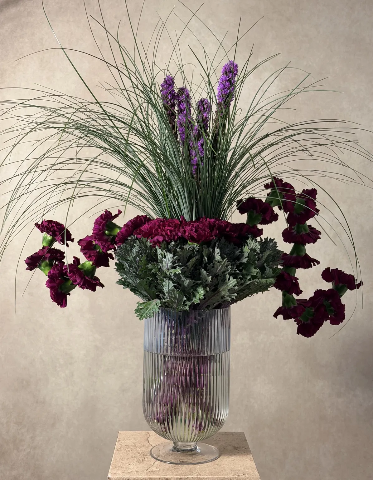 Purple liatris and grasses rising above deep magenta carnations and kale in a ribbed glass vessel