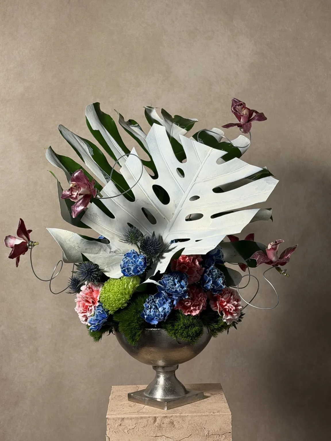 Front of the silver-vessel piece: a white-painted monstera leaf over blue hydrangea, carnations and floating orchids in a pewter urn