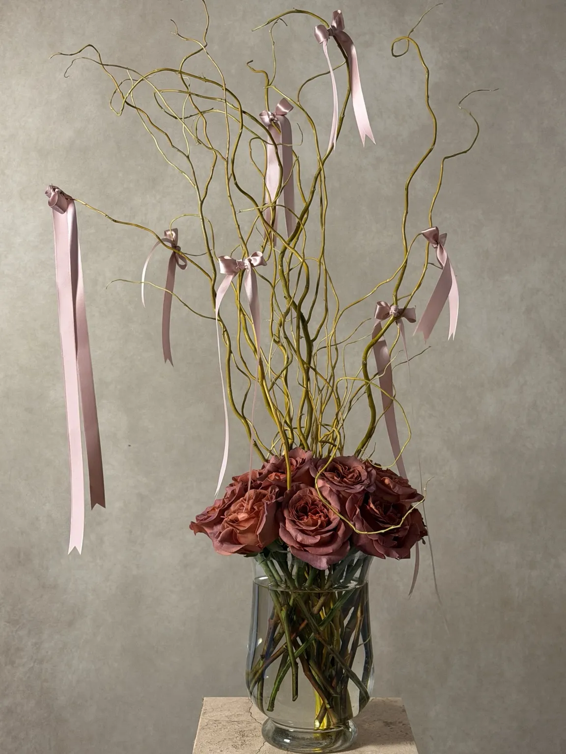 Tall curly willow branches hung with pale pink ribbons above dusty mauve roses in a glass vase