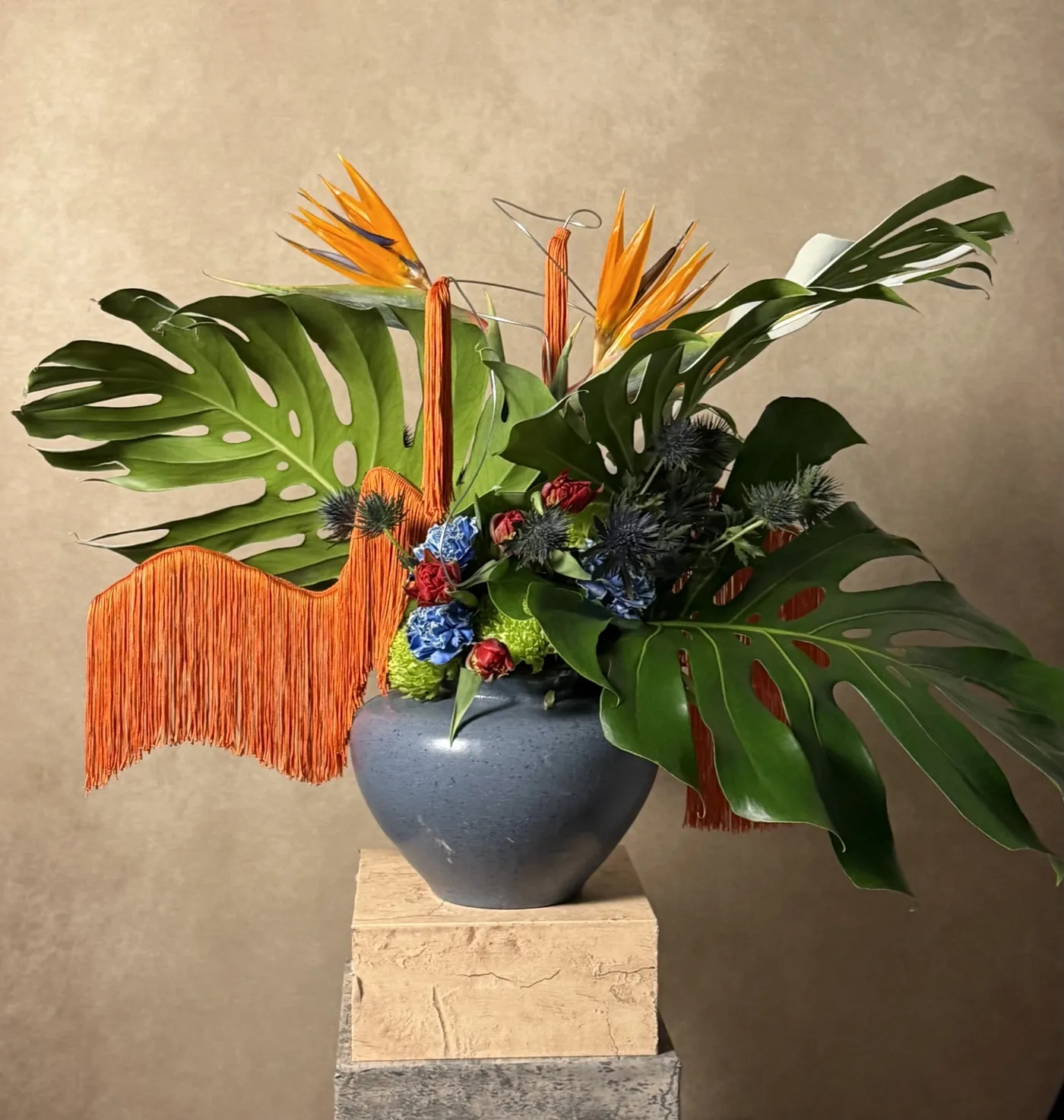 Sculptural arrangement of monstera leaves, bird of paradise, blue carnations and orange fringe in a slate-blue vessel on a stone plinth
