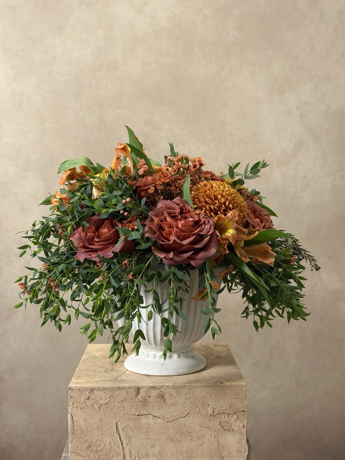 Autumn arrangement of rust roses, orange blooms and loose greenery in a white pedestal vase